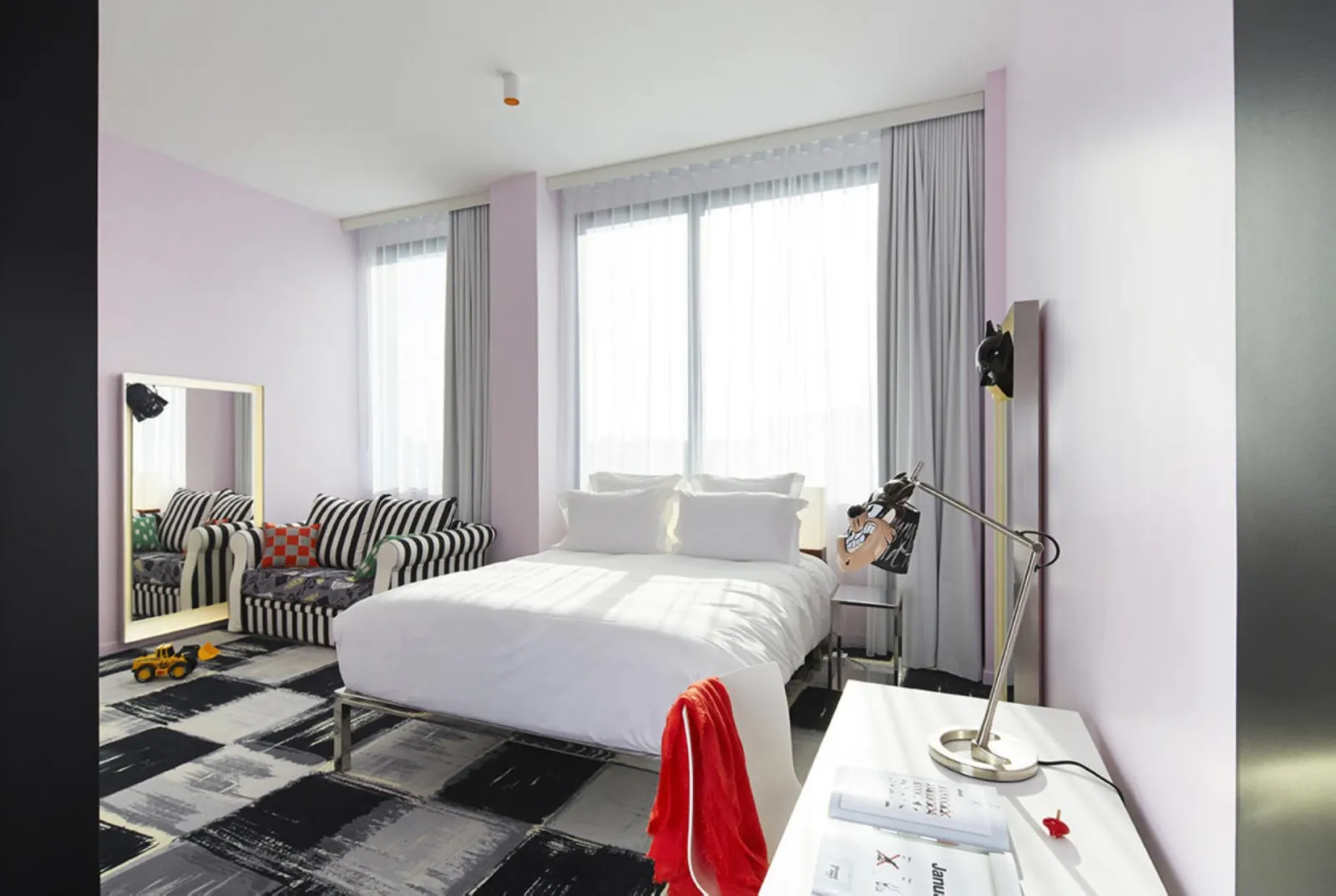 Modern bedroom at Mama Shelter Bordeaux featuring a white bed, gold-framed mirror, black and white striped armchairs, large windows with gray curtains, and contemporary black and red décor.