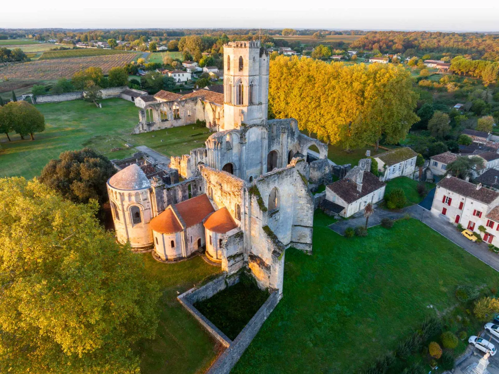 Aerial view of the Abbey of La Sauve-Majeure near Bordeaux, displaying the ruins of the medieval monastery with its Romanesque bell tower, churches with red tile roofs, surrounded by greenery and autumn fields.