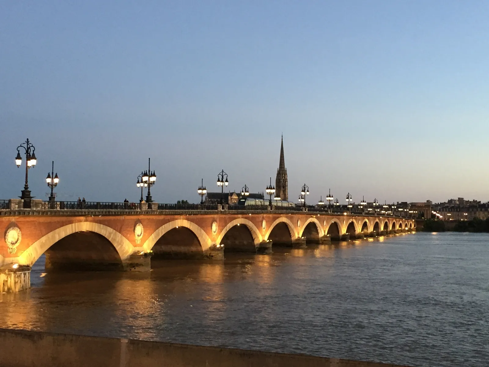 The Stone Bridge of Bordeaux lit up at dusk, spanning the Garonne River with its golden stone arches, Saint-Michel Cathedral visible in the background