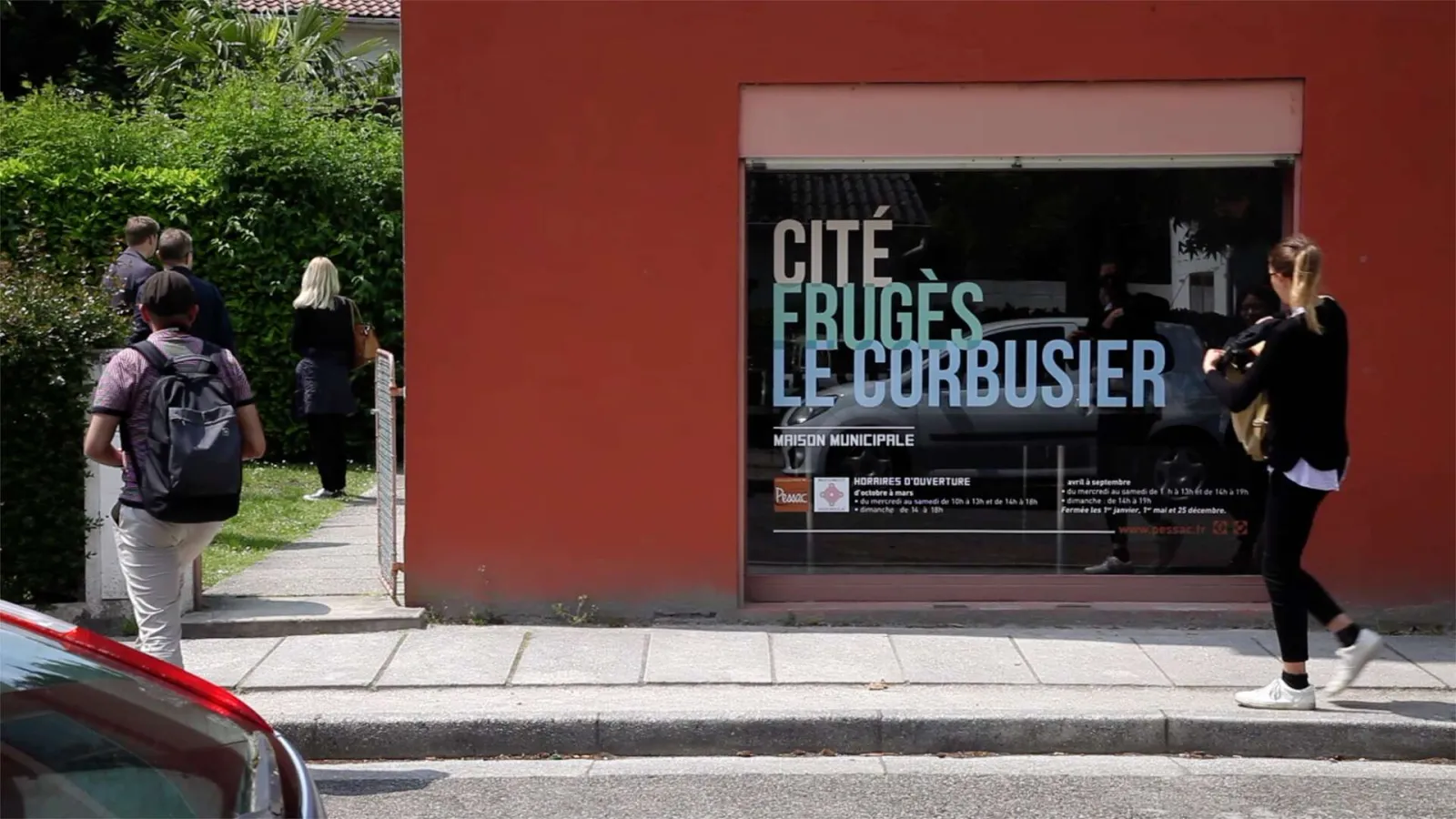 Red facade of the Municipal House at Cité Frugès Le Corbusier in Pessac, with a window display showcasing a temporary exhibition, visitors observing the architecture.