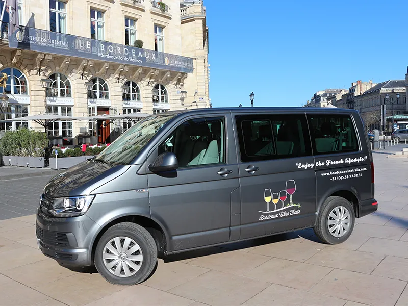 A gray minibus parked in a Bordeaux square, decorated with wine glass decals and text reading 'Enjoy the French lifestyle' and 'Bordeaux Wine Trails' on its sides, in front of a historic building.
