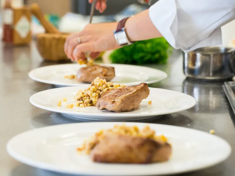 Cooking workshop at Château Kirwan in Bordeaux: a chef in white jacket plates three dishes of grilled meat with corn kernels in a professional kitchen.