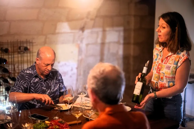 A woman presents a wine bottle during a tasting in Bordeaux, facing guests seated around a table with glasses and food.