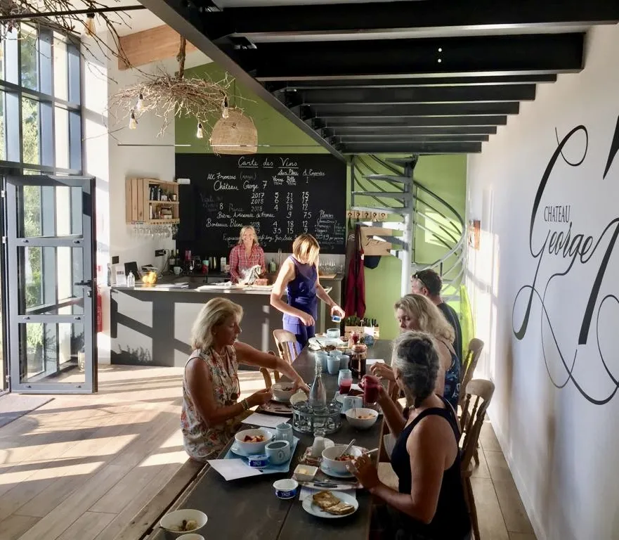 Modern restaurant or wine bar interior with guests seated at a black table, staff at the counter in background, geometric green and white decor, and handwritten menu on blackboard.