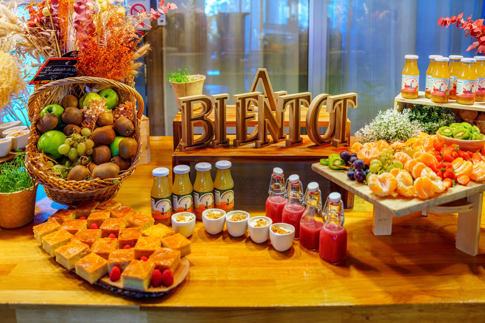 Fresh fruit and juice buffet display featuring a fruit basket, fruit pastries, juice glasses, and pastis bottles at Novotel Bordeaux Lac, decorated with golden autumn elements and BIENTÔT lettering.