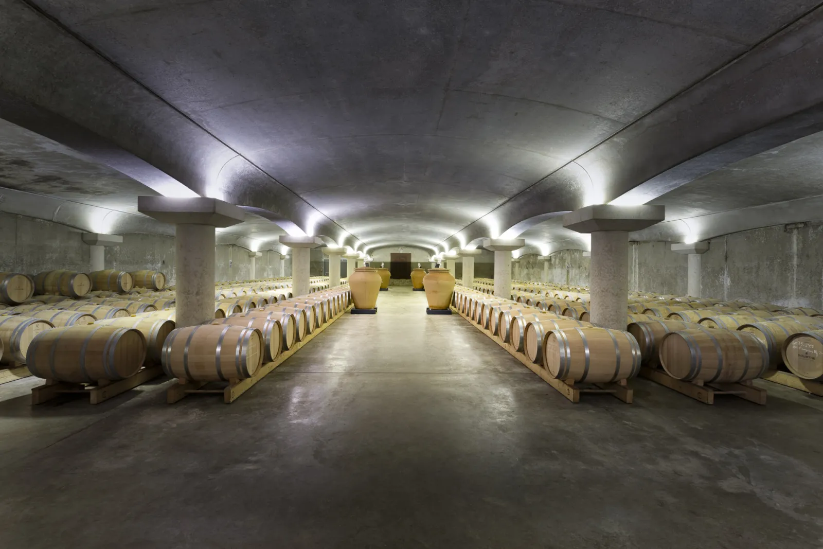 Underground cellar at Château La Garde in Bordeaux featuring symmetrical rows of wooden barrels on either side of a central illuminated aisle, concrete columns, and two large golden amphorae at the end