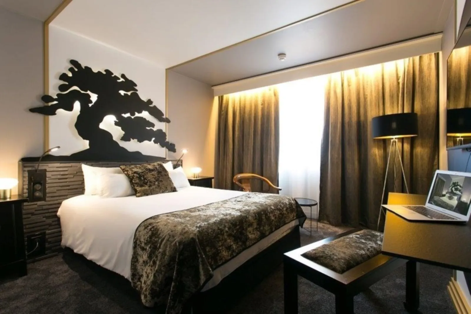Modern hotel room featuring a queen-size bed, black dragon wall art, golden curtains, desk with laptop, and contemporary black and gold furnishings.