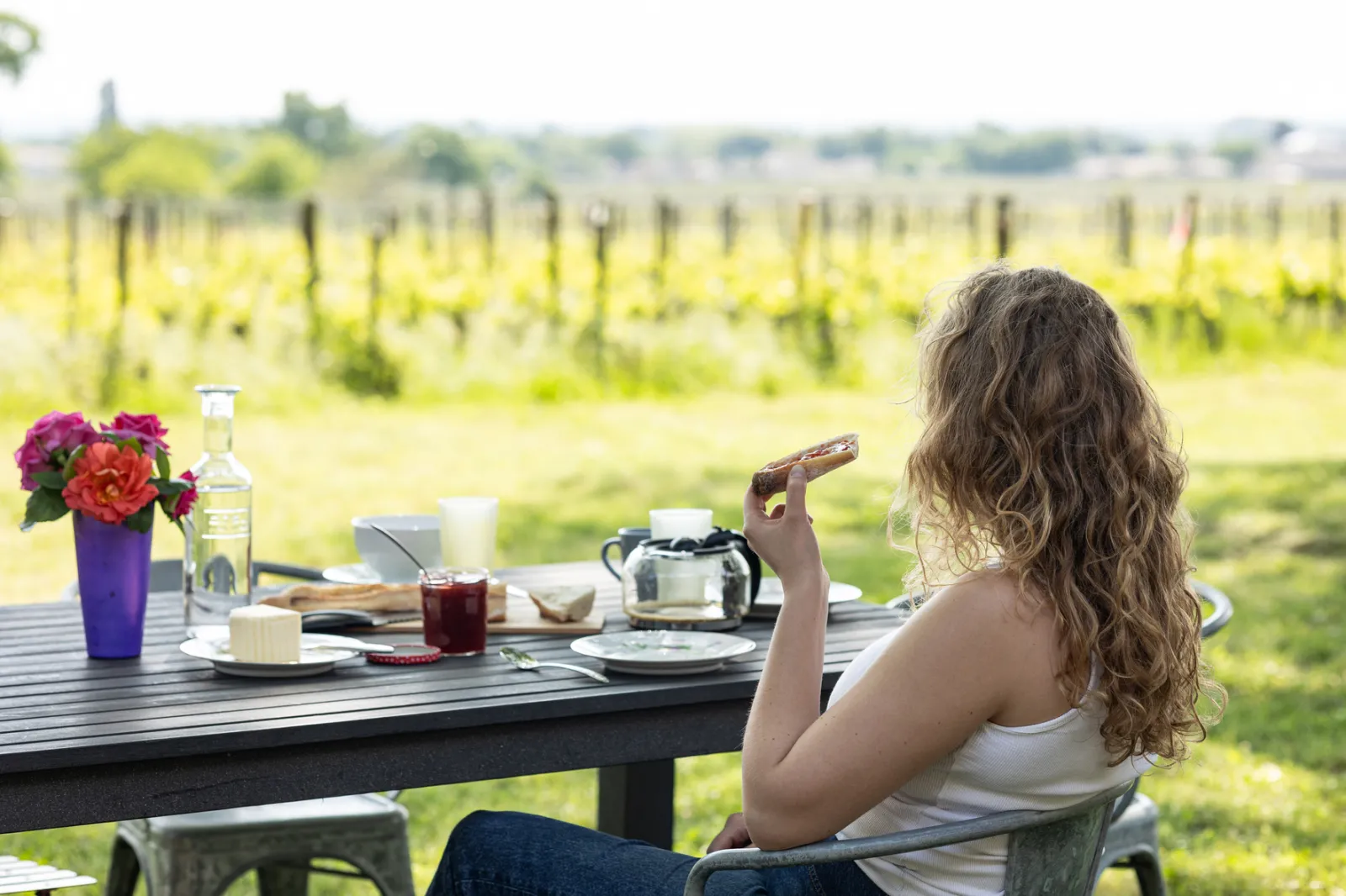 A long-haired woman enjoys a cigar at a picnic table overlooking Bordeaux vineyards, with flowers, wine and dining accessories.
