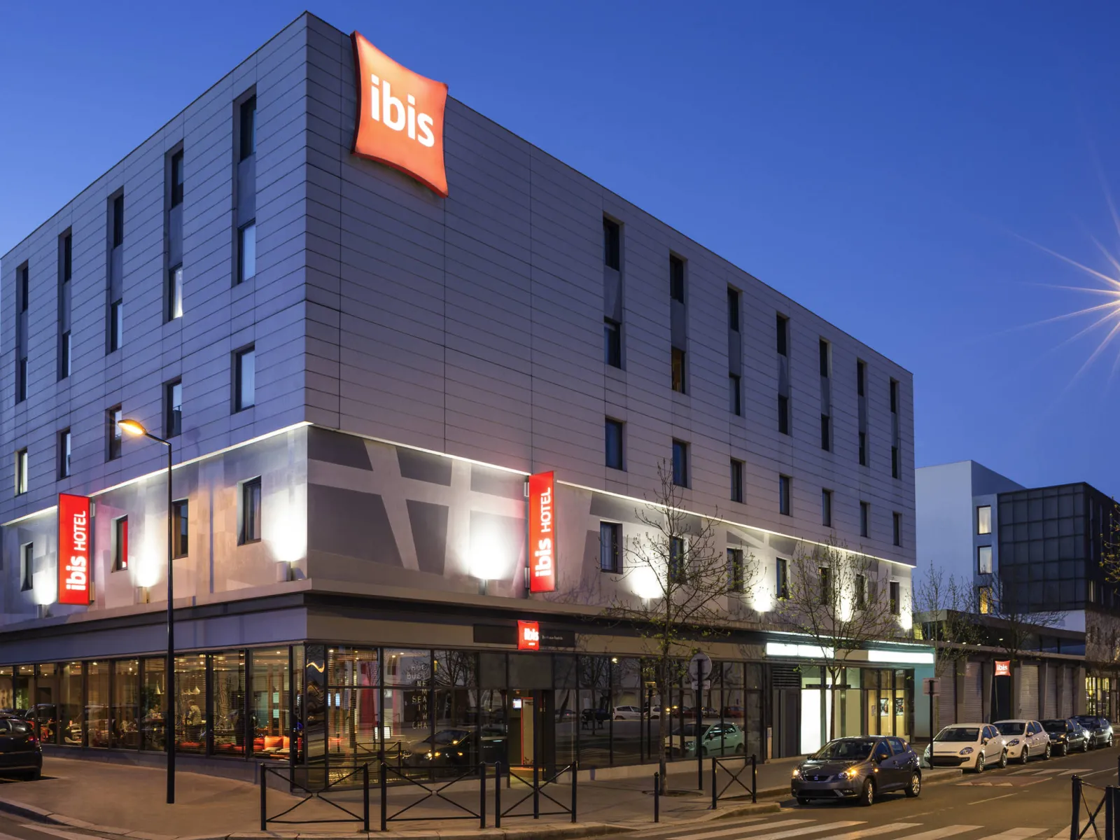 Modern facade of Ibis Bordeaux Centre Bastide hotel at night, featuring contemporary gray and white architecture, illuminated red signage, and parked cars in front