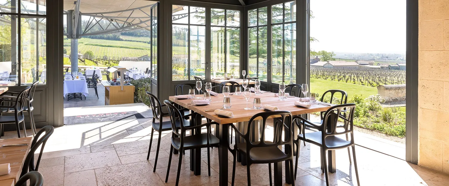 Modern restaurant dining room with floor-to-ceiling windows overlooking Bordeaux vineyards, wooden table with black designer chairs, exterior terrace visible, abundant natural light.