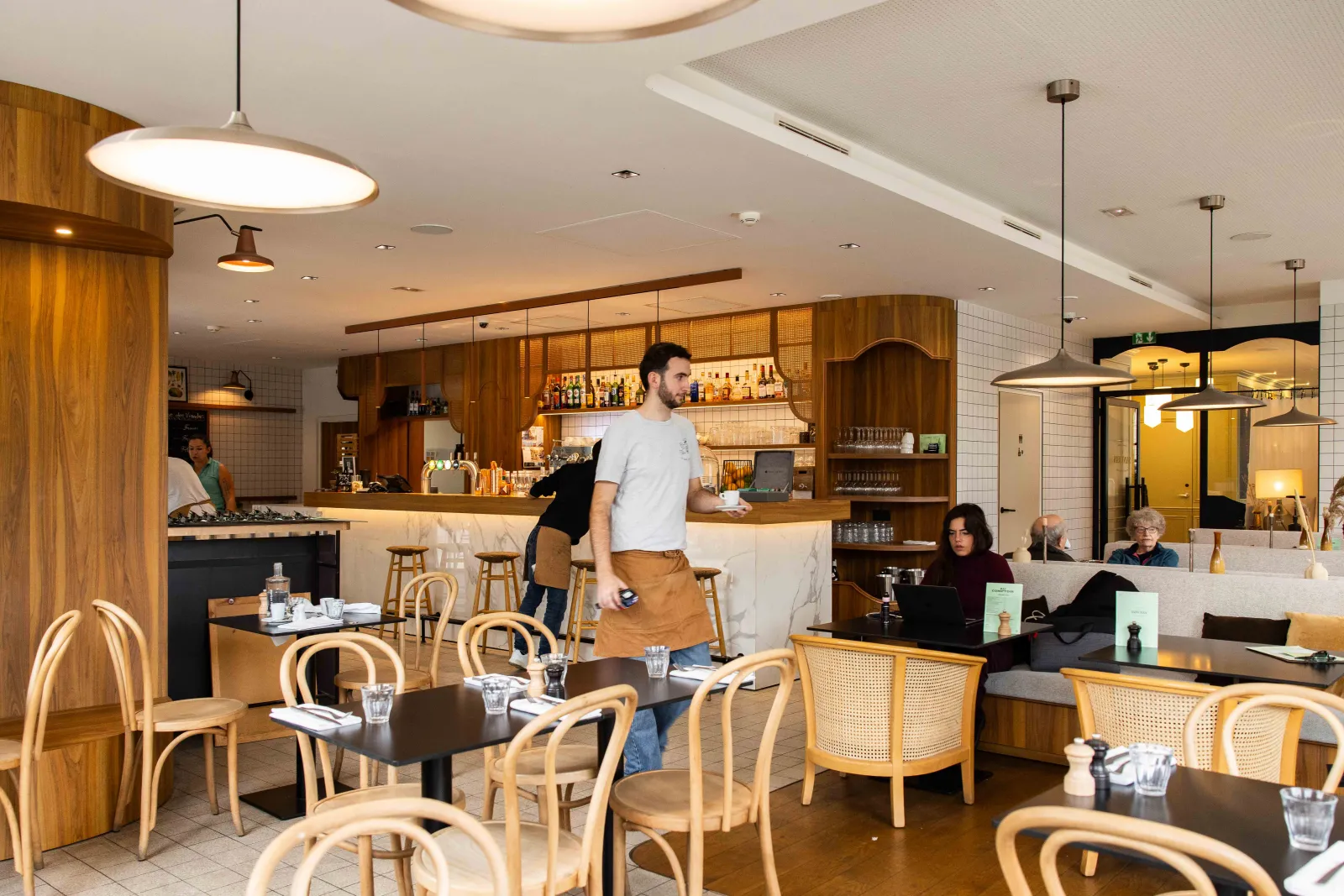 Interior of Bistrot Le Saint-Jean in Bordeaux featuring wooden counter, rattan chairs, seated guests, staff members, modern lighting, and warm atmosphere.