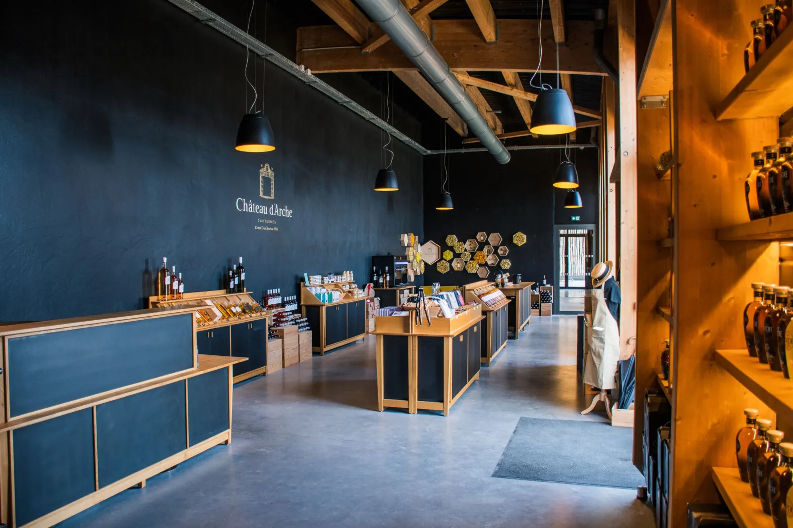 Modern interior of Château d'Arche shop in Bordeaux featuring dark blue walls, wooden counters, orange-lit shelves, black and yellow pendant lights, and displays of wine products and accessories.