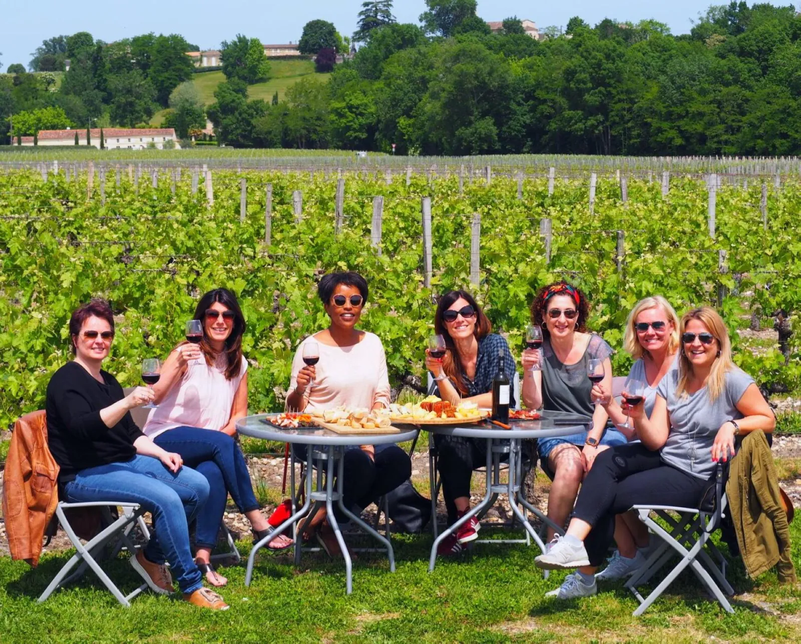 Seven women seated around a table with wine glasses and charcuterie, in a Bordeaux vineyard with buildings and trees in the background