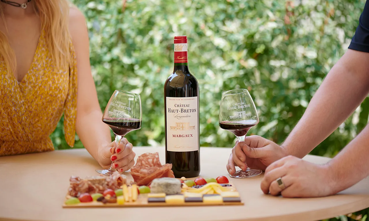 Two people enjoy a red wine from Château Haut-Breton Larigaudière Margaux in Bordeaux, with filled wine glasses and a charcuterie and cheese board, in a lush green garden.