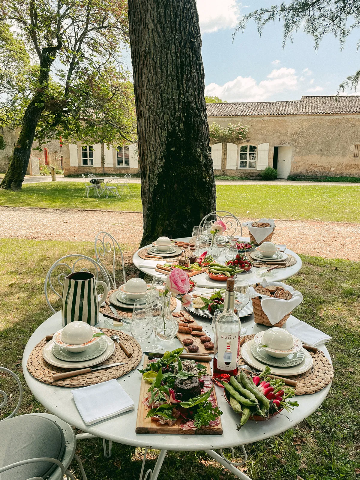 An elegantly set outdoor dining table beneath a large tree at Château de Cérons, featuring fine tableware, fresh flowers, and gourmet dishes with the stone château facade in the background.