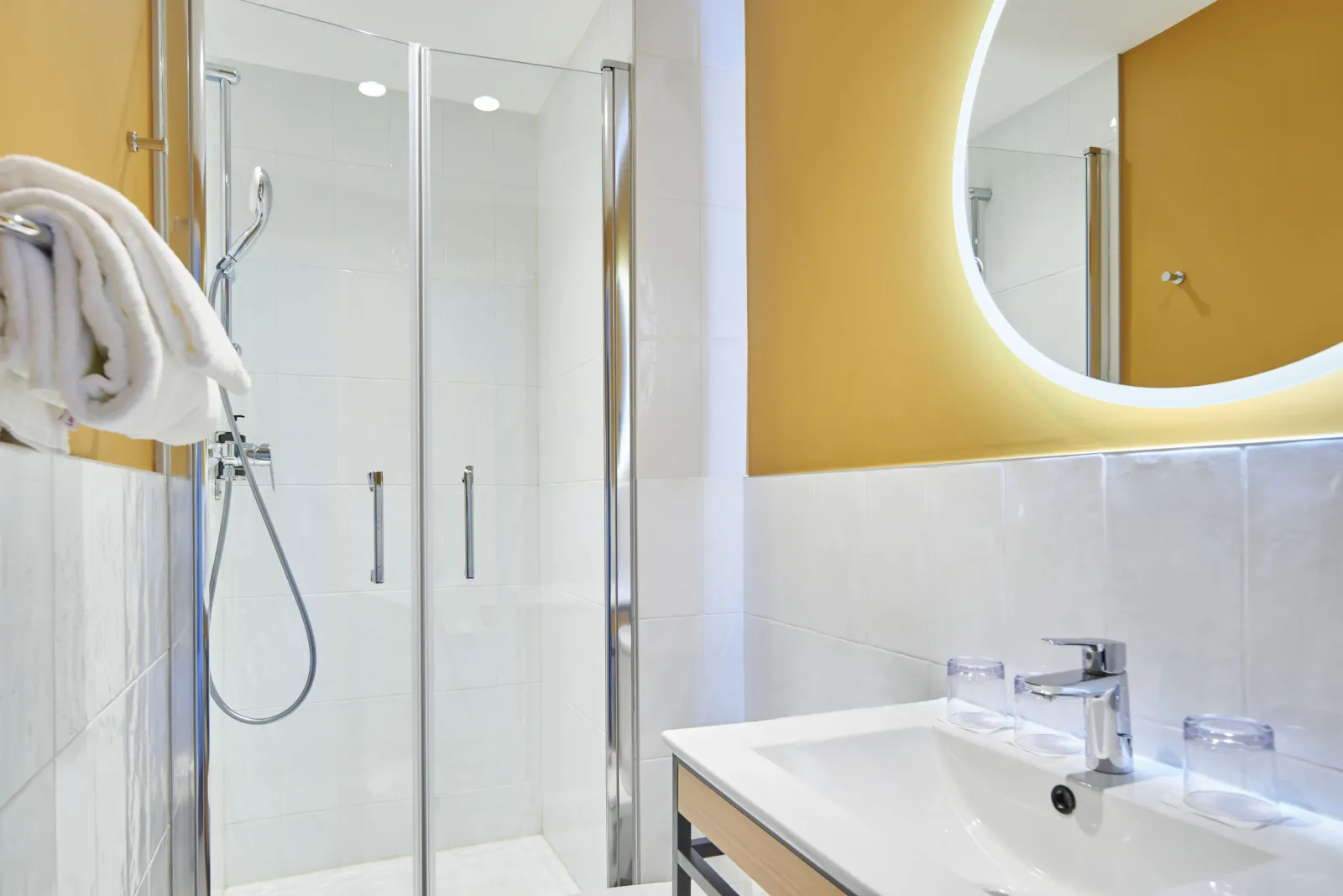 Modern bathroom with mustard yellow and white walls, walk-in shower with glass partition, illuminated round mirror, white sink and white towels