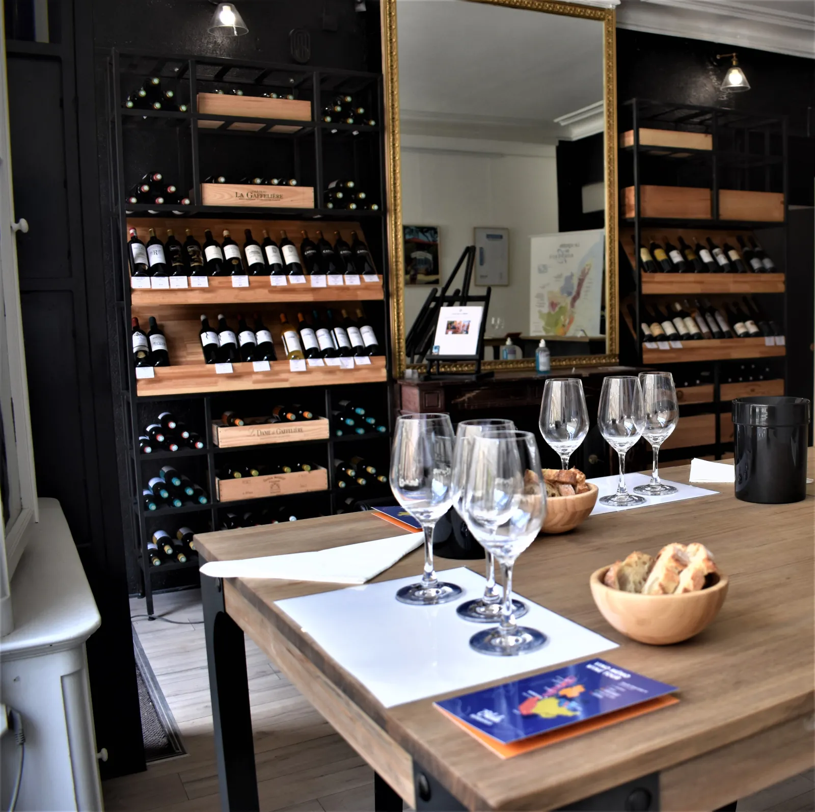Wine shop interior in Bordeaux featuring a wooden tasting counter with wine glasses, snacks in a basket, and extensive bottle display on wooden shelves against black and gold decorated walls.