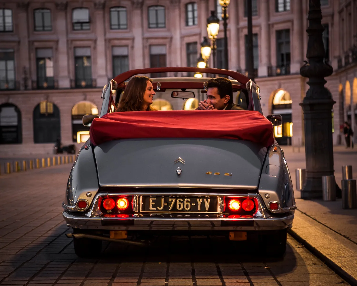 Couple in a vintage grey convertible with red soft top driving through a historic Bordeaux square, pale pink illuminated facades in the background