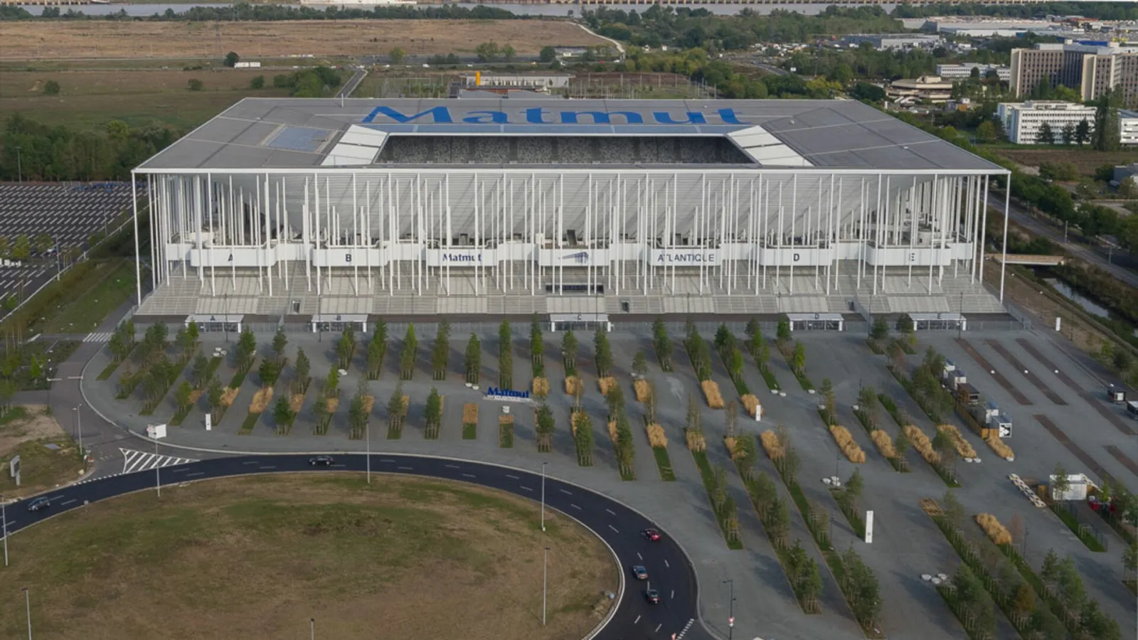Aerial view of the Matmut Atlantique stadium in Bordeaux, featuring a modern glass-fronted structure with tree-lined parking lot and surrounding urban landscape.