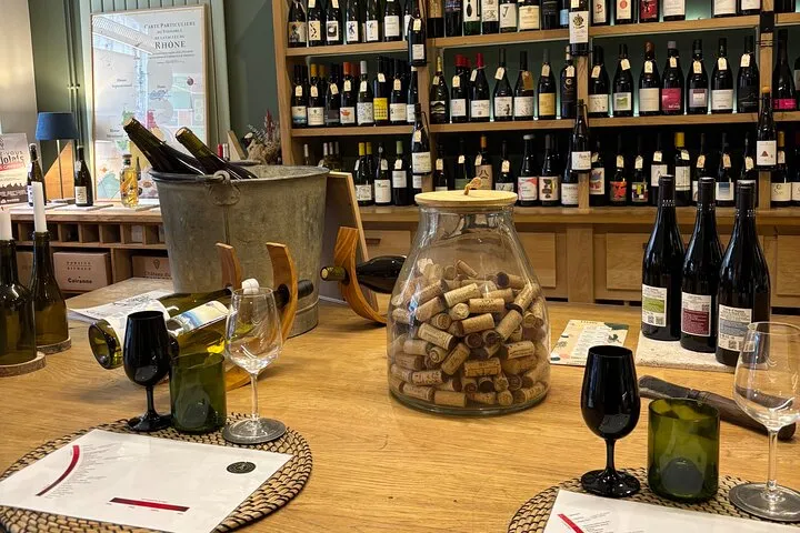 Interior of a Bordeaux wine cellar with a tasting counter displaying bottles, glasses, an ice bucket, and a jar filled with wine corks, facing shelves stocked with wines.