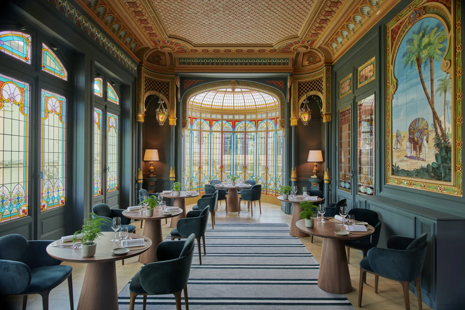 Elegant dining room at Château Hôtel Grand Barrail in Bordeaux, featuring ornate gilded coffered ceiling, stained glass windows, light wood round tables, dark blue chairs, and orientalist wall paintings.