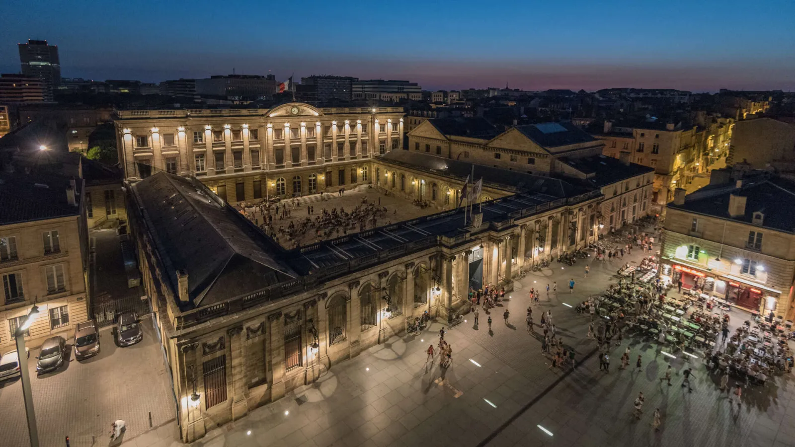 Night view of Bordeaux's illuminated Palais Rohan with its classical stone facades, surrounded by historic buildings and busy public squares.