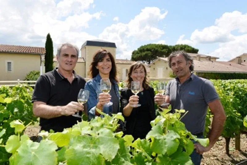 Four people holding white wine glasses in a vineyard at Château Haut-Bergeron in Bordeaux, with wine estate buildings in the background.