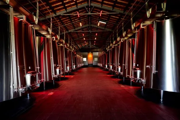 Interior of Maison Cardinale winery in Bordeaux, featuring a long corridor lined with red and stainless steel fermentation tanks, red floor, and exposed wooden ceiling.
