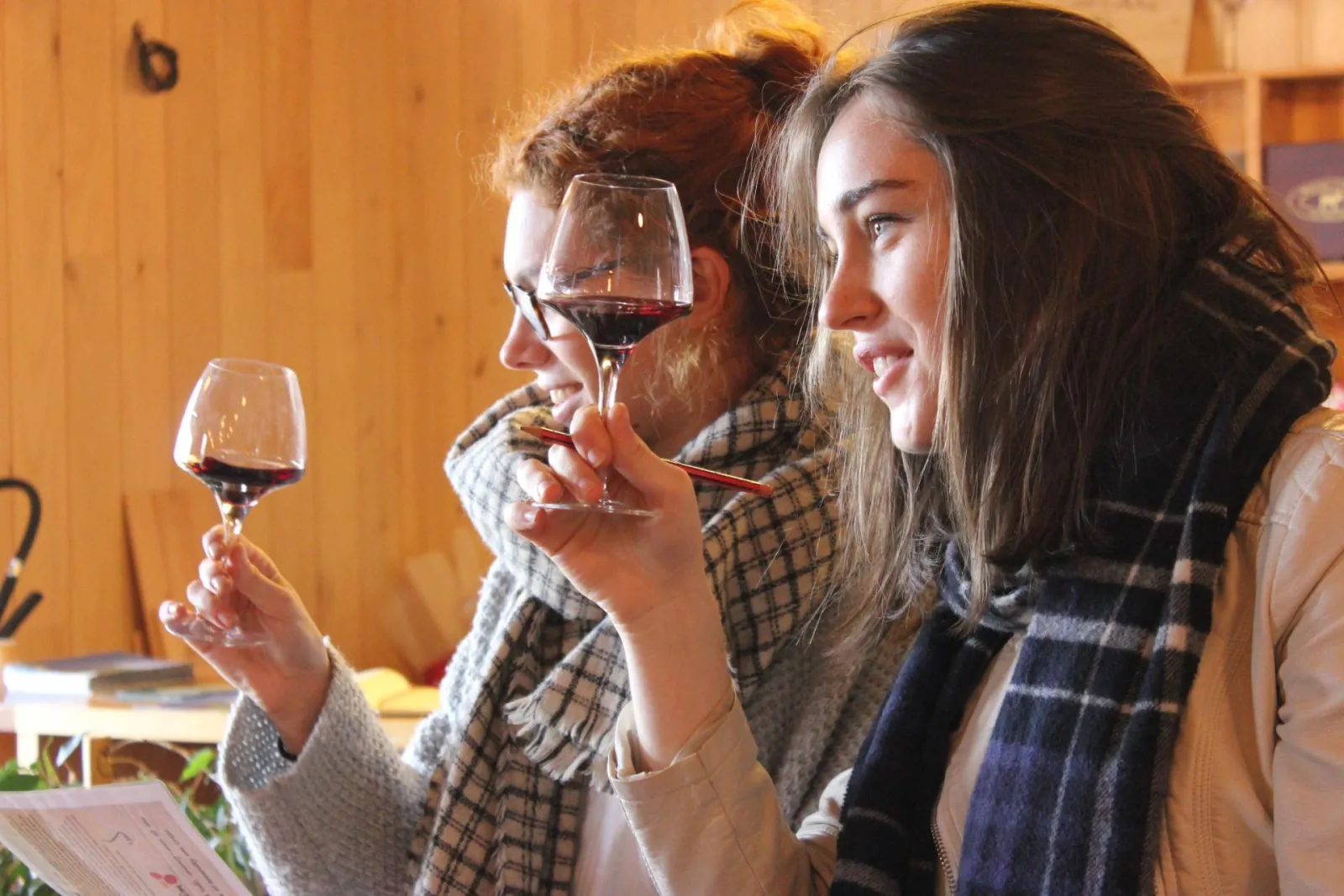Two people taste red wine in a wooden cellar at Château Pas de l'Âne in Bordeaux, carefully examining their wine glasses.