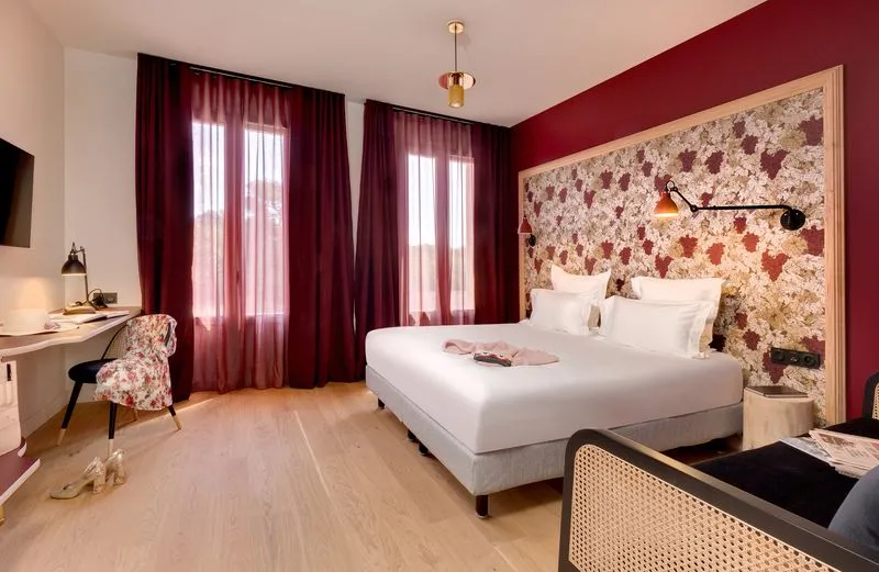 Elegant hotel bedroom featuring a queen-size bed with white linens, burgundy walls and matching curtains, floral wallpaper headboard, desk, patterned chair, and light wood flooring.