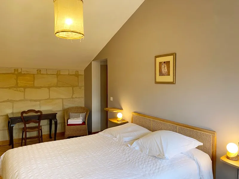 Elegant bedroom featuring a queen-size bed with white linens, rattan headboard, soft beige walls, sloped ceiling, designer lighting fixtures, antique desk, wicker chair and exposed stone wall.