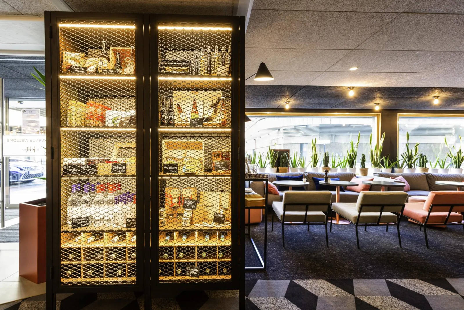 Modern lounge interior featuring an illuminated wine display case, colorful seating, and a view of Bordeaux in the background.