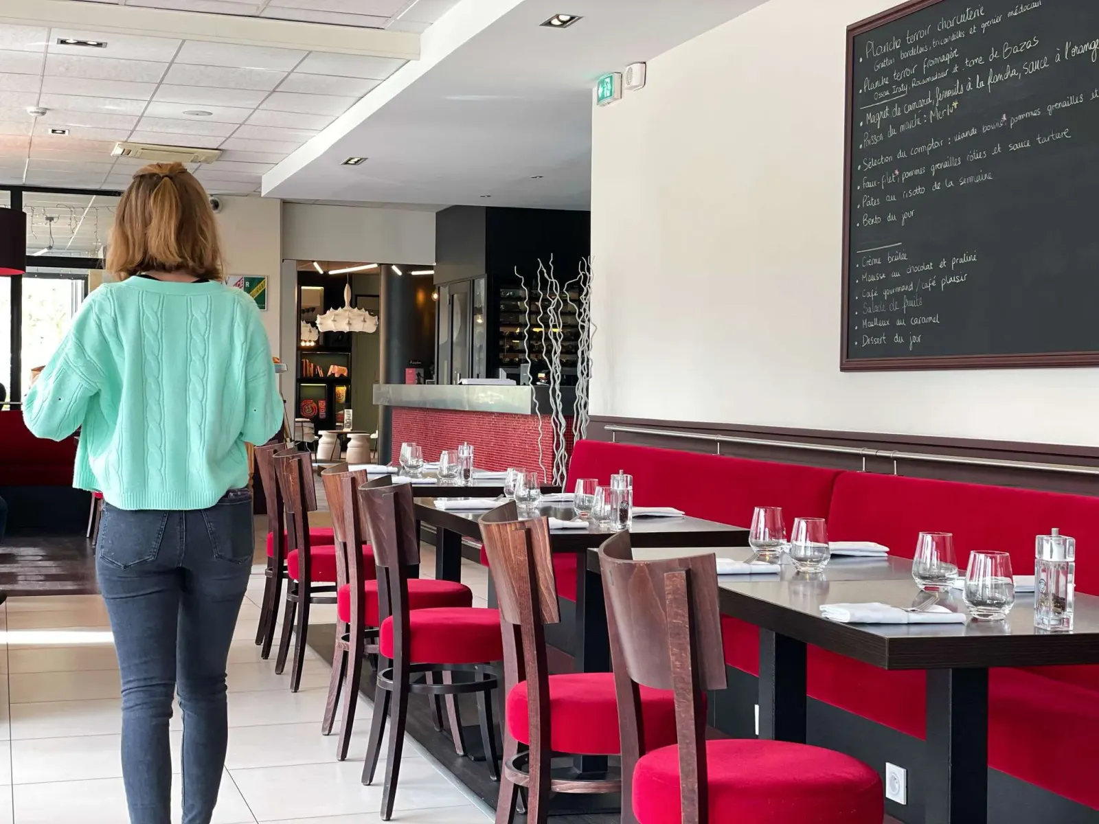 Interior of a Bordeaux restaurant showing a woman in a turquoise shirt standing before a row of tables with red banquettes, wooden chairs, water glasses, and a chalk menu board on the wall.