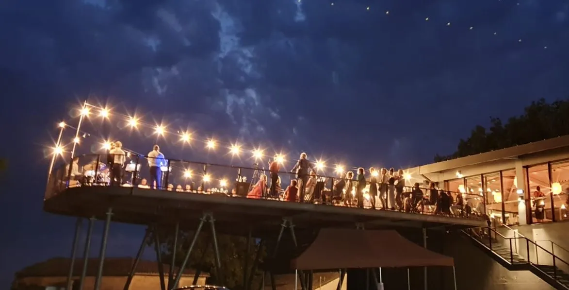 An illuminated platform with string lights overhanging a modern building, with a crowd of people gathered at dusk in Bordeaux at Château Réaut.