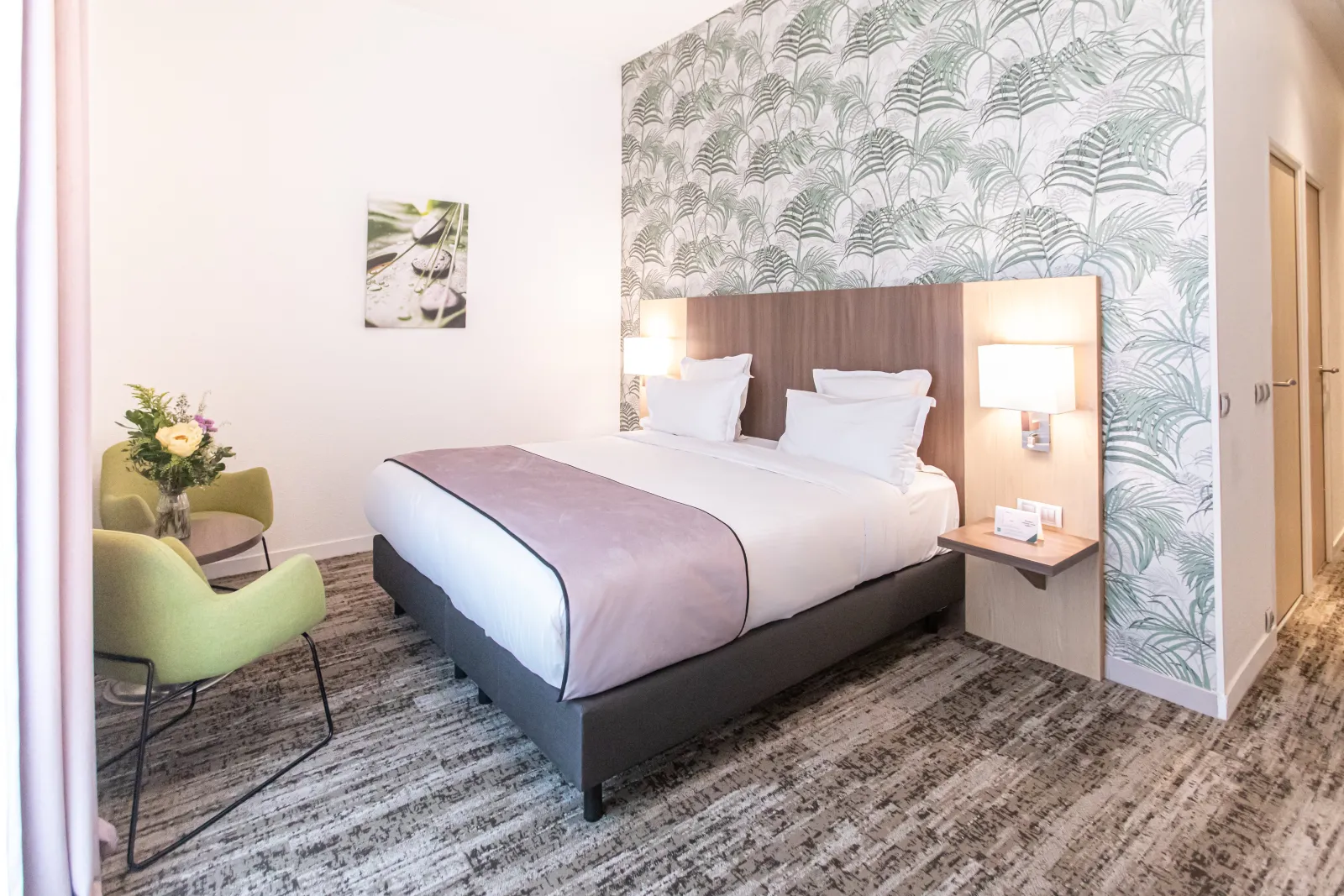 Modern hotel room featuring a queen-size bed with taupe headboard, botanical gray-green patterned wallpaper, lime green armchair, side table with flower arrangement, and contemporary furnishings.