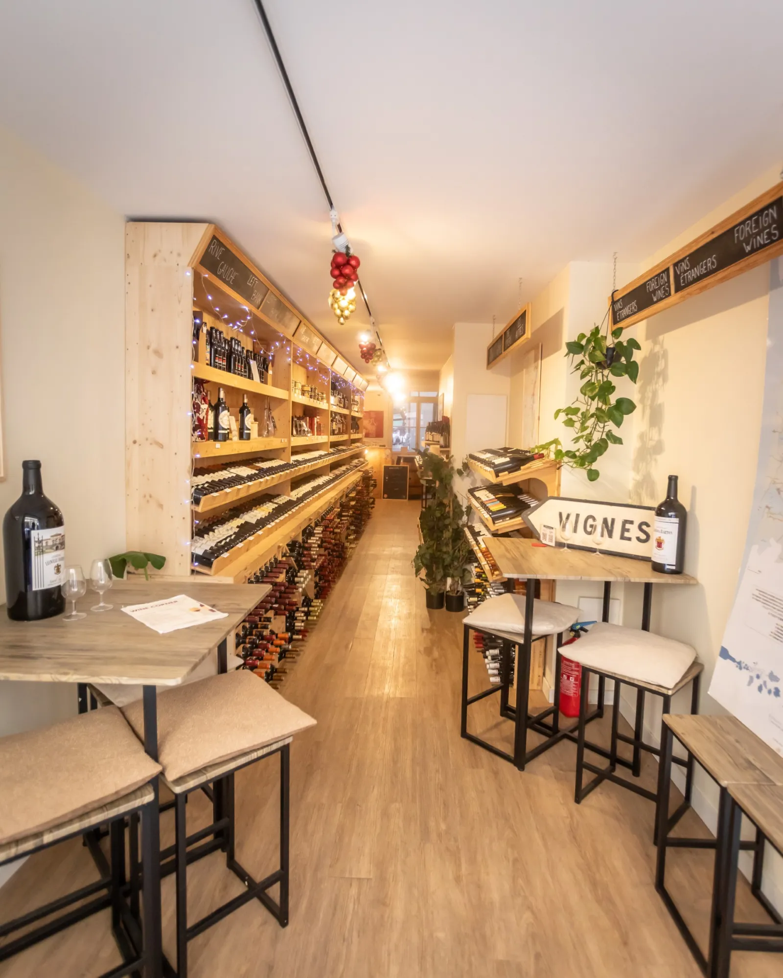 Interior of Wine Corner in Bordeaux, a narrow wine bar with wine bottle shelves lining the walls, high tables with stools, warm lighting and green plants.