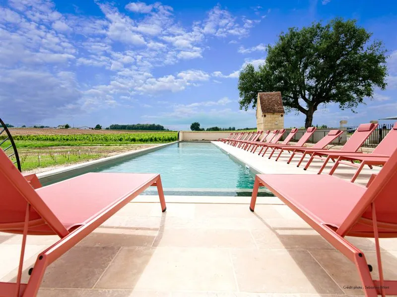 Outdoor swimming pool with pink lounge chairs surrounding a turquoise pool, overlooking vineyards and an old tower at Château Fage, Bordeaux.