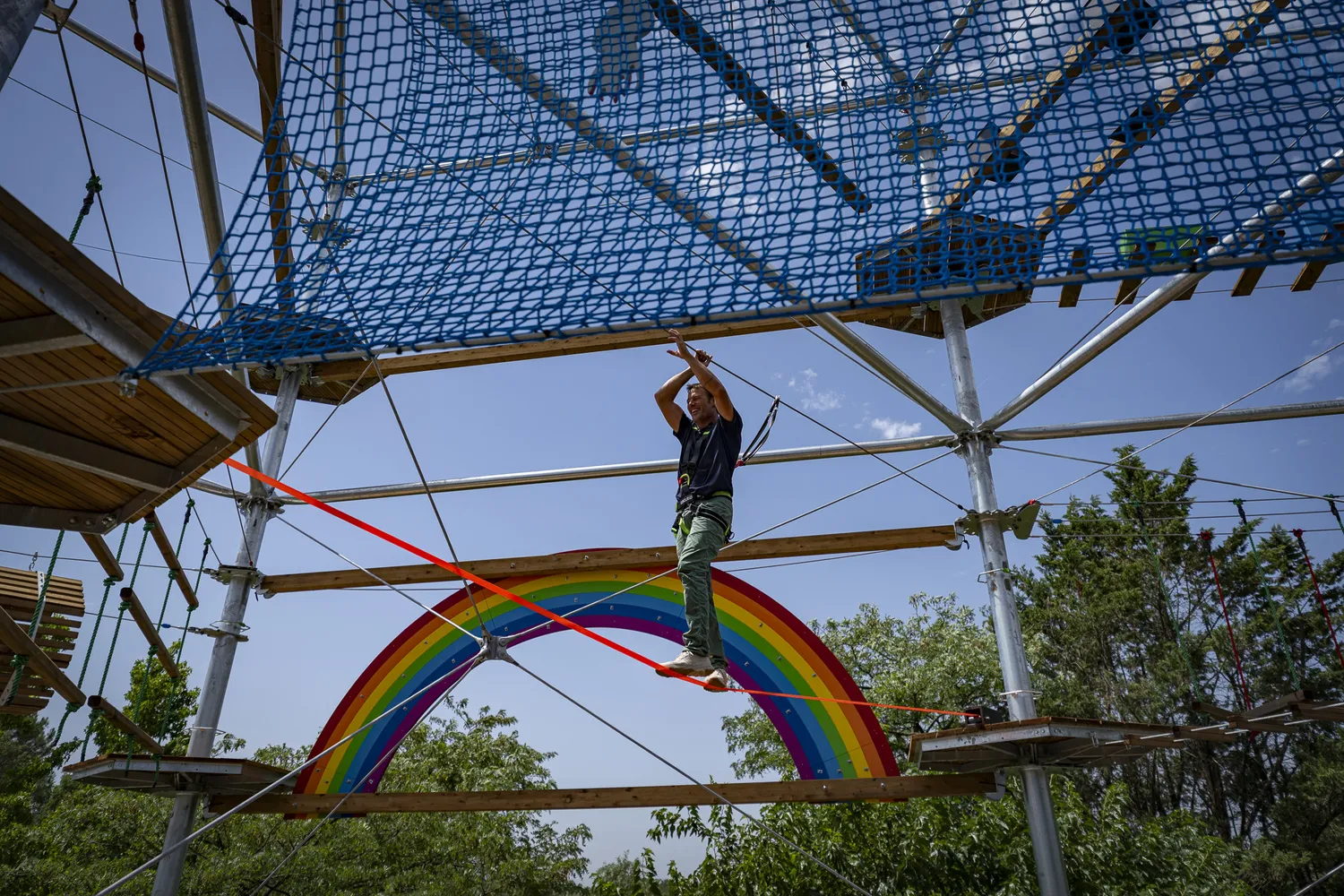 A participant zip-lining through a high rope adventure course at Cimes de Bordeaux Pessac, crossing a rope and net structure with a colorful rainbow in the background