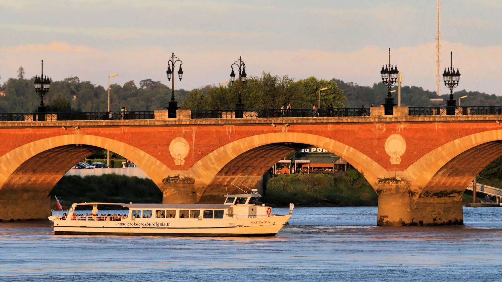 Bordeaux's Stone Bridge with its red brick arches spanning the Garonne River, a cruise boat passing under the bridge at sunset