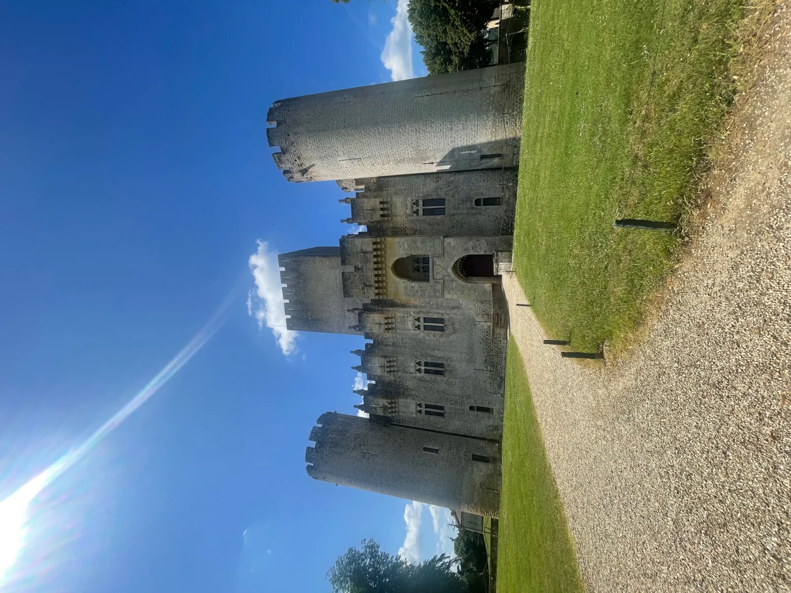 View of Roquetaillade Castle near Bordeaux, a medieval fortress featuring massive towers with crenellations, photographed against a clear blue sky with green lawn in the foreground.