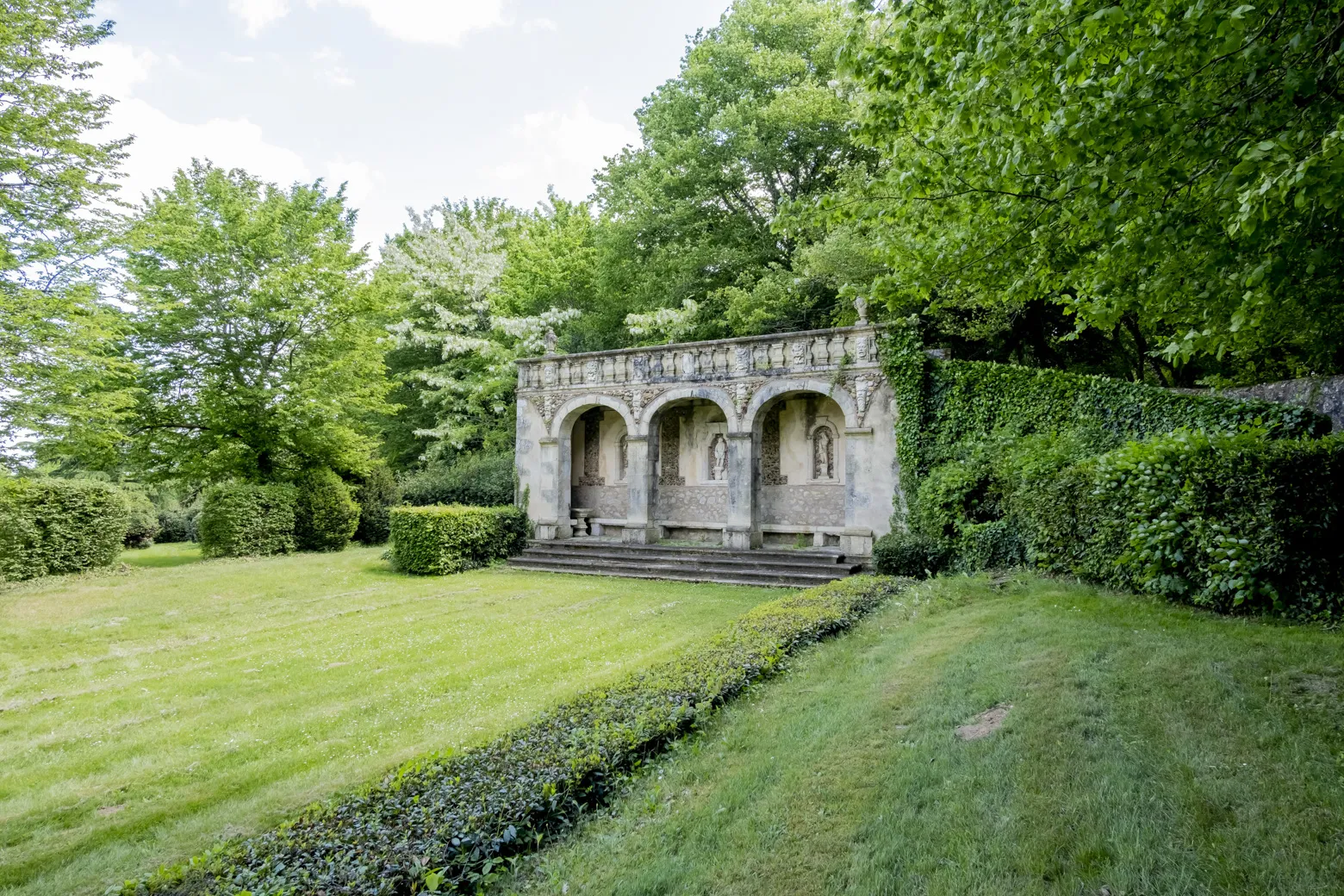 Stone pavilion with three arches in a landscaped garden at Château de Malle in Bordeaux, surrounded by lush greenery and hedges.