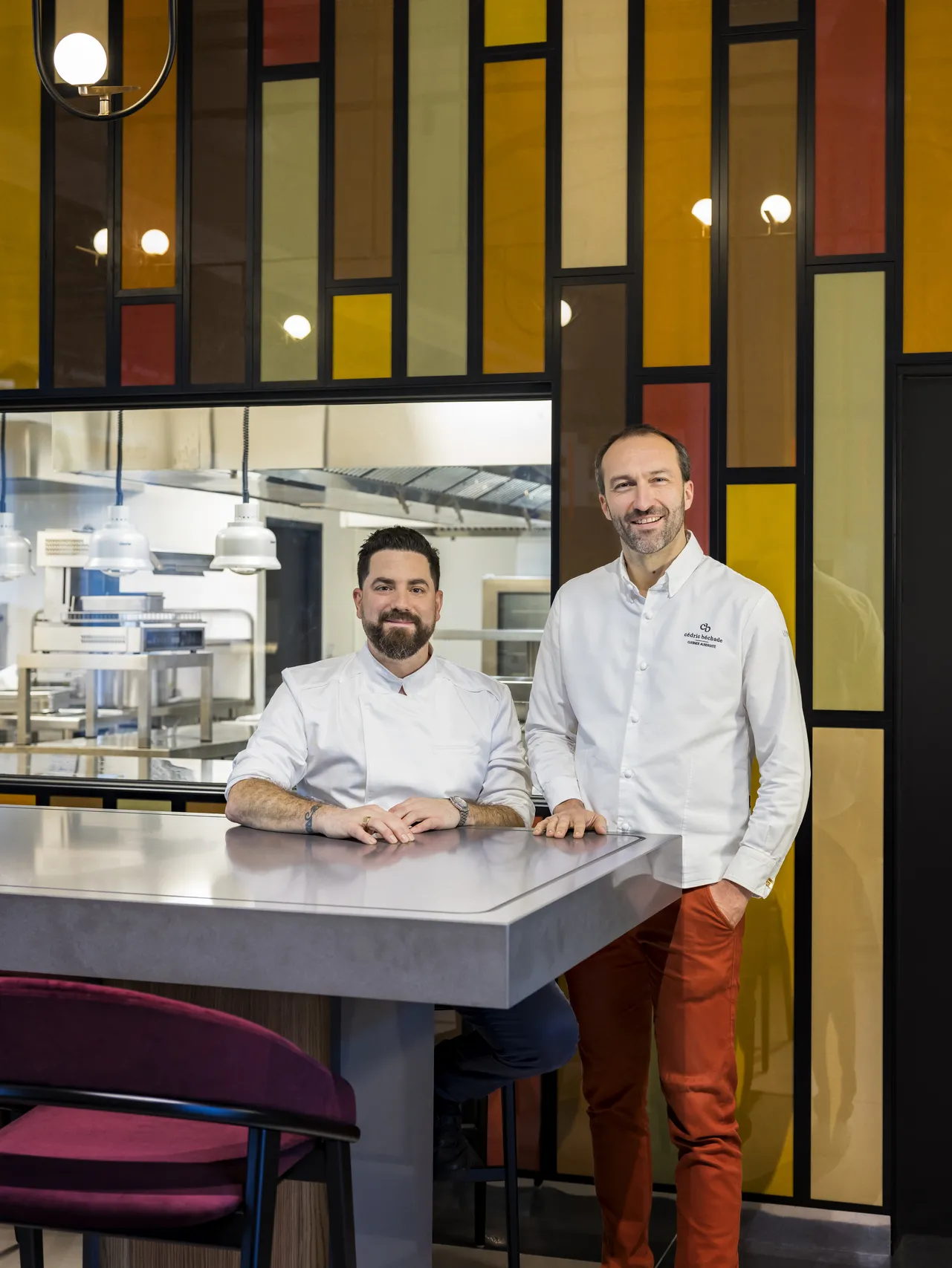Two chefs in white chef's coats stand at the counter of a modern restaurant in Bordeaux, with an open kitchen visible behind them and a colorful geometric design in the background.
