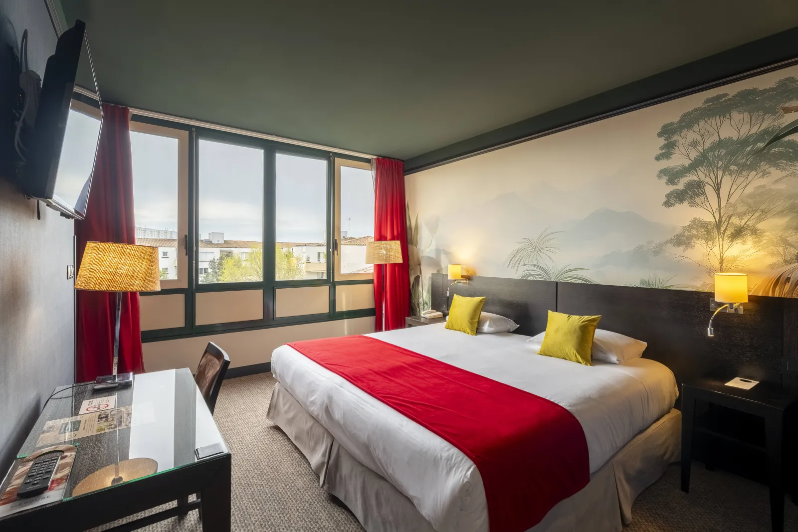 Modern hotel room featuring a king-size bed with white linens and red throw, yellow pillows, large windows overlooking Bordeaux, tropical wall mural, warm yellow lighting and contemporary furnishings.