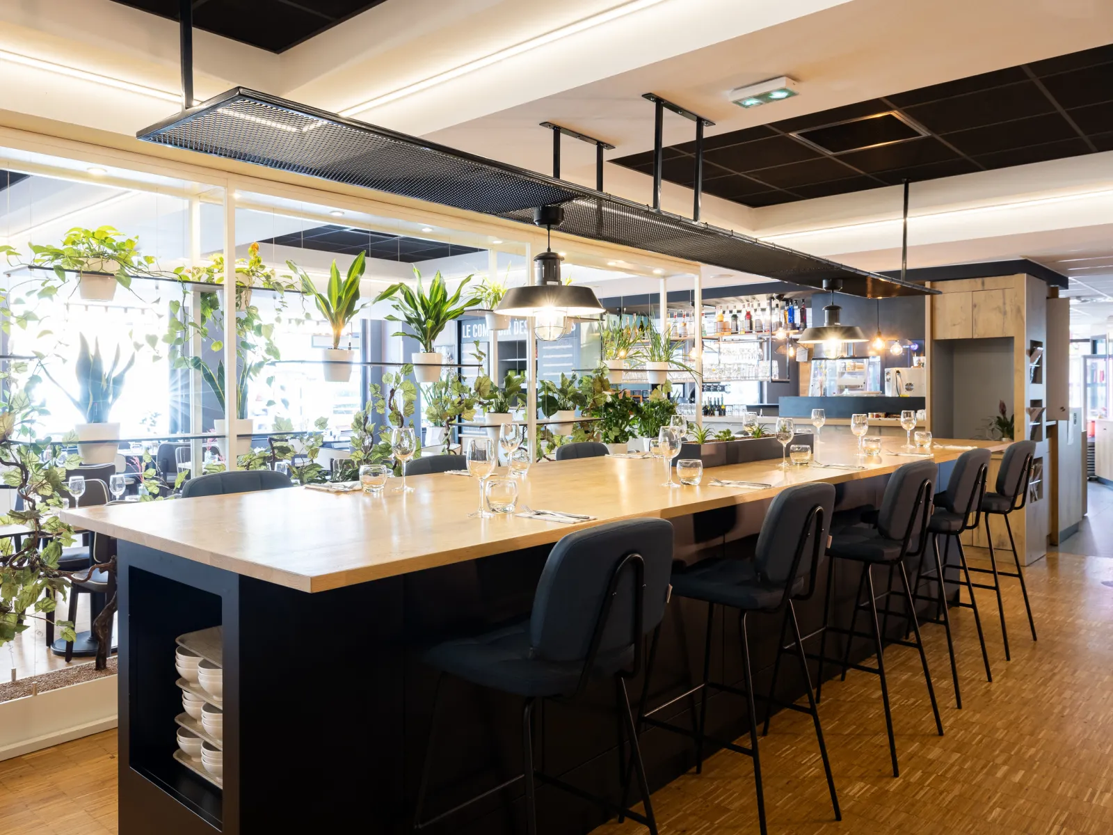 Modern interior of Bistrot de Mériadeck in Bordeaux featuring a large wooden counter, black bar stools, green plants, pendant lighting, and street-facing windows.