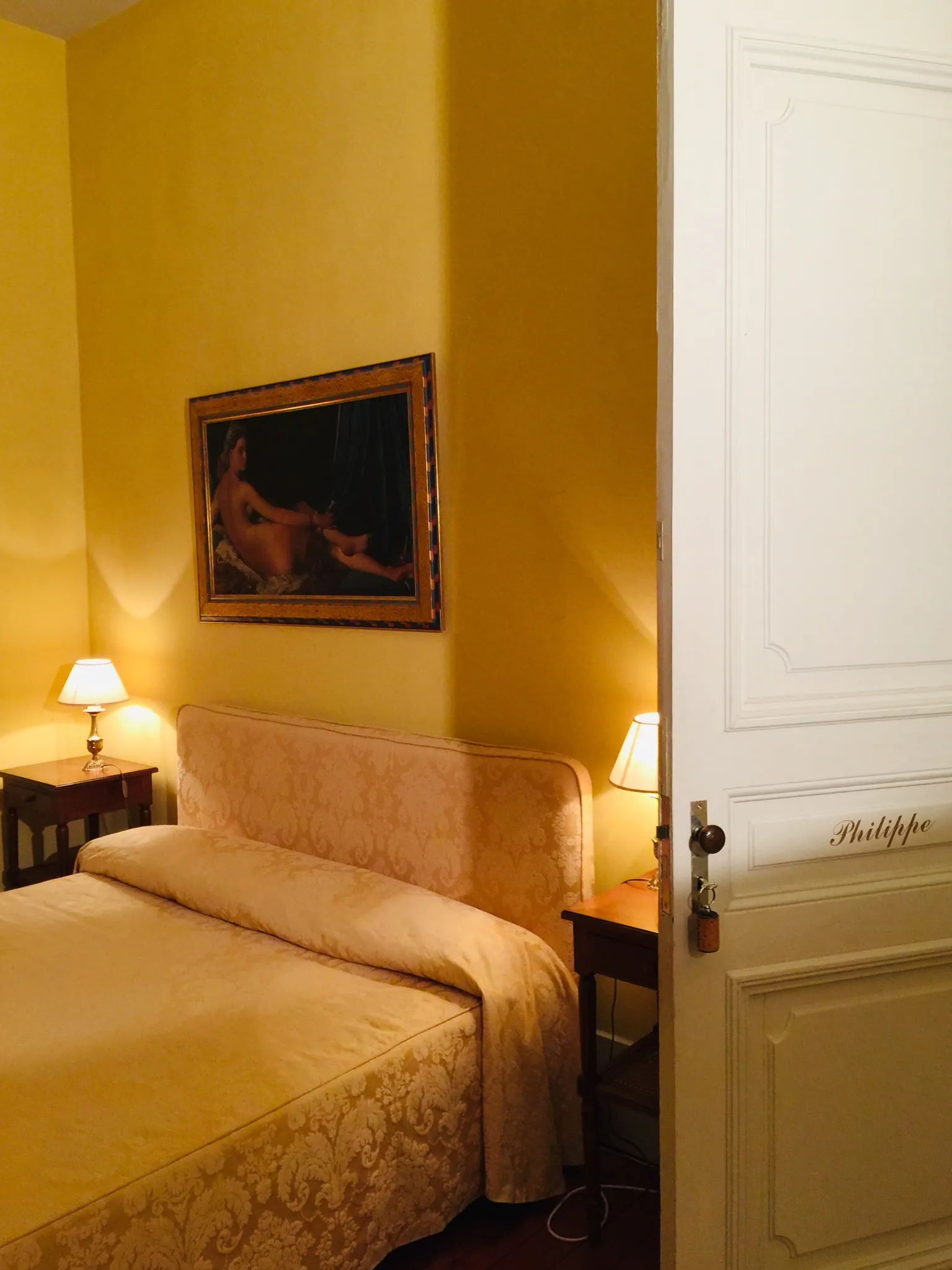 Elegant guest bedroom with golden yellow walls, a bed with beige linens, two lit bedside lamps, a framed painting above the bed, and a white door on the right