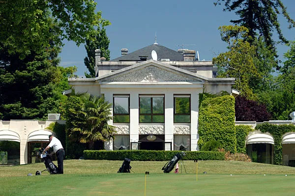 Clubhouse of Golf de Margaux near Bordeaux featuring white neoclassical facade with columns, grey roof, and golfers practicing on the green lawn.