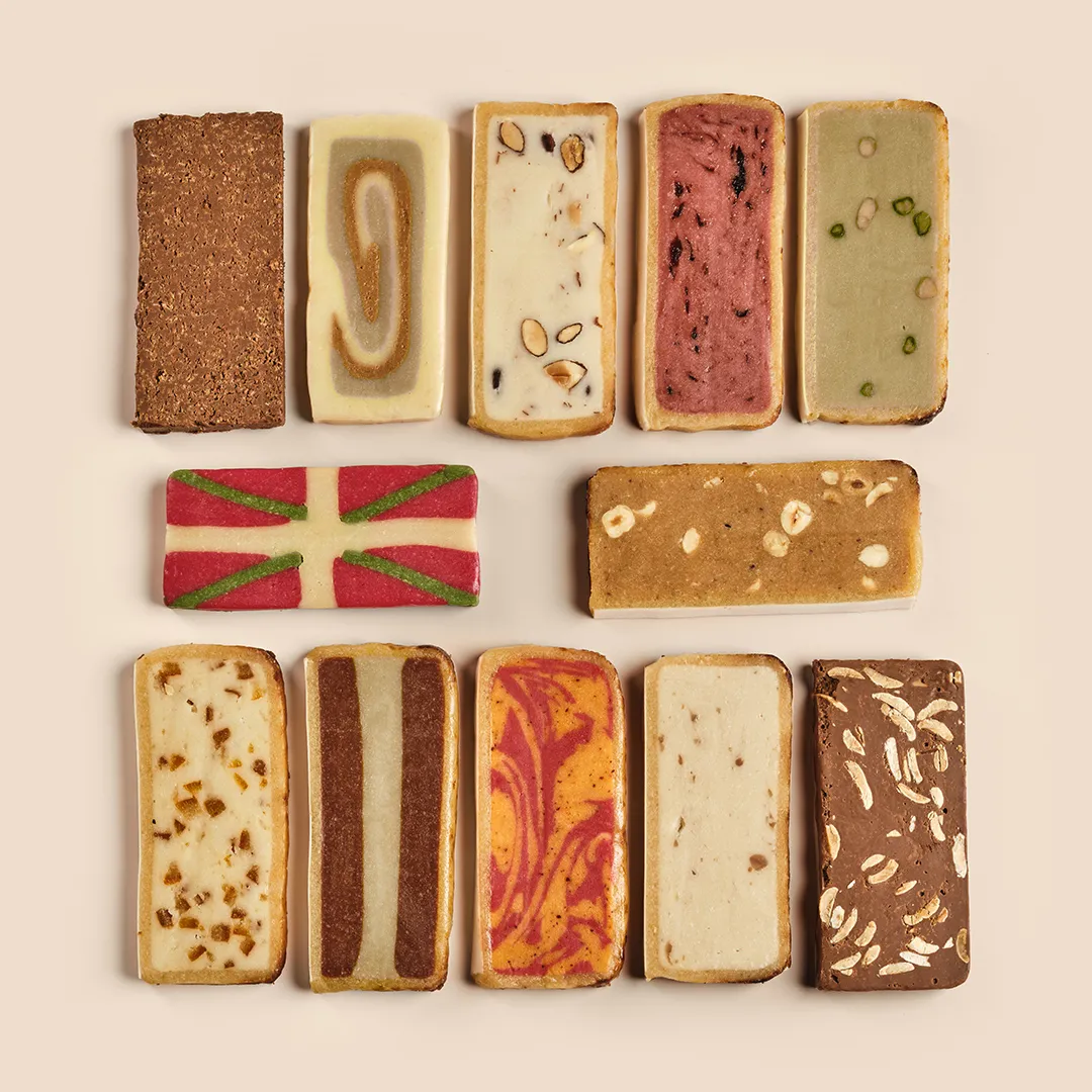 Thirteen artisanal small cakes from Maison Pariès in Bordeaux, featuring various flavors and toppings including chocolate, nuts, almond paste, and traditional colorful designs.