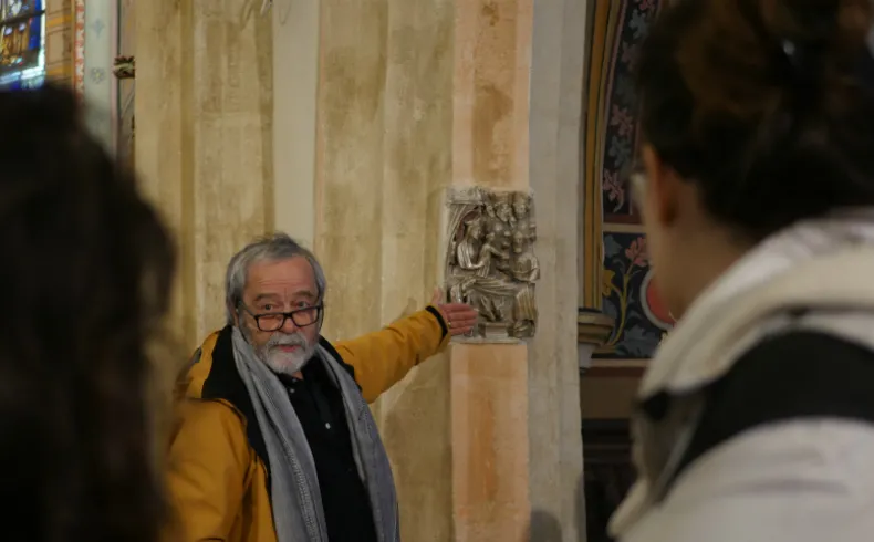 An elderly man wearing glasses and a mustard-yellow jacket with a gray scarf points to a stone ornamental sculpture on a pillar during a guided tour in the Old Lormont district of Bordeaux.