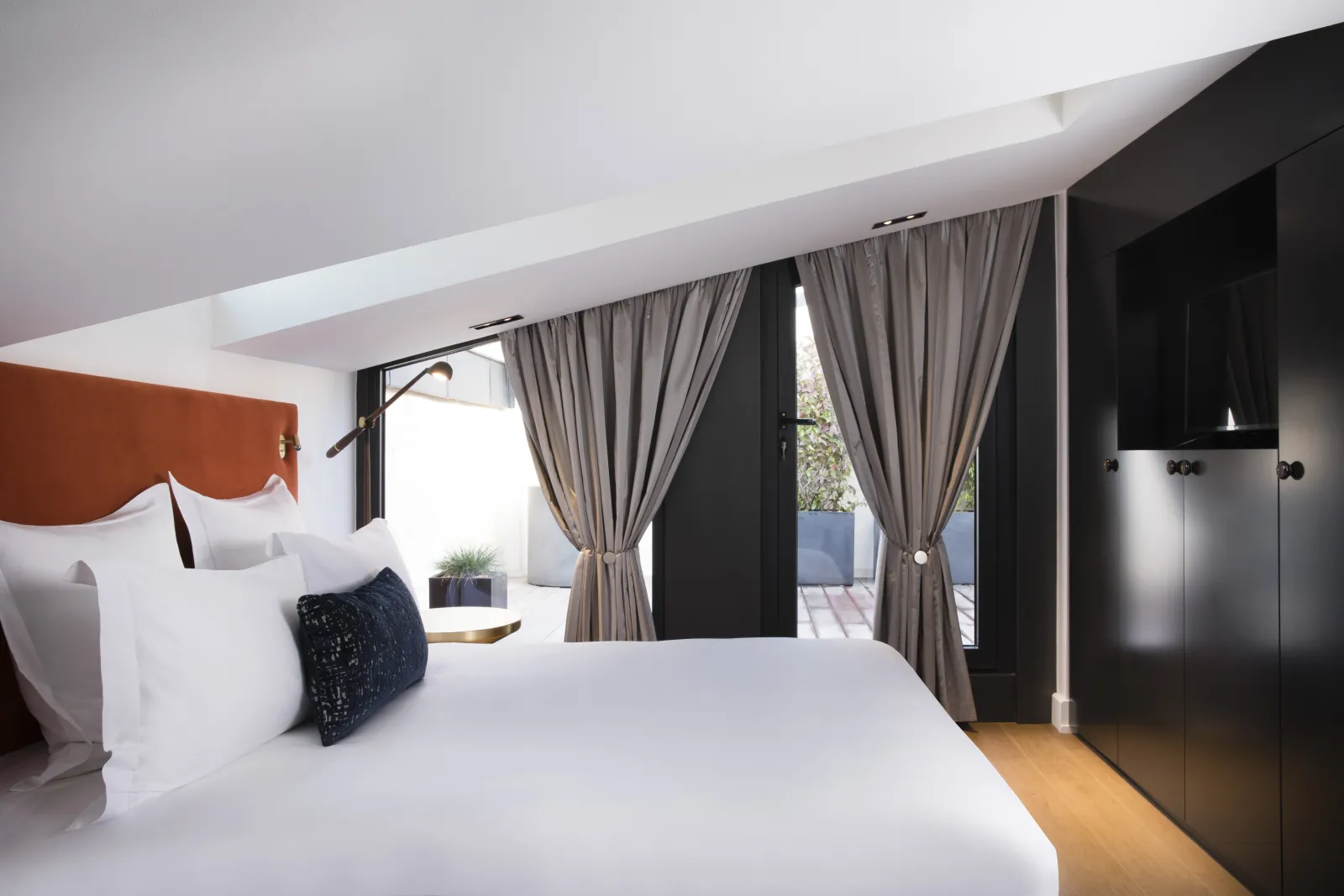 Luxury modern bedroom at Hôtel Cardinal Bordeaux featuring a king-size bed with terracotta headboard, floor-to-ceiling windows opening onto a terrace, beige curtains, contemporary black furniture, and white ceiling with exposed beams.