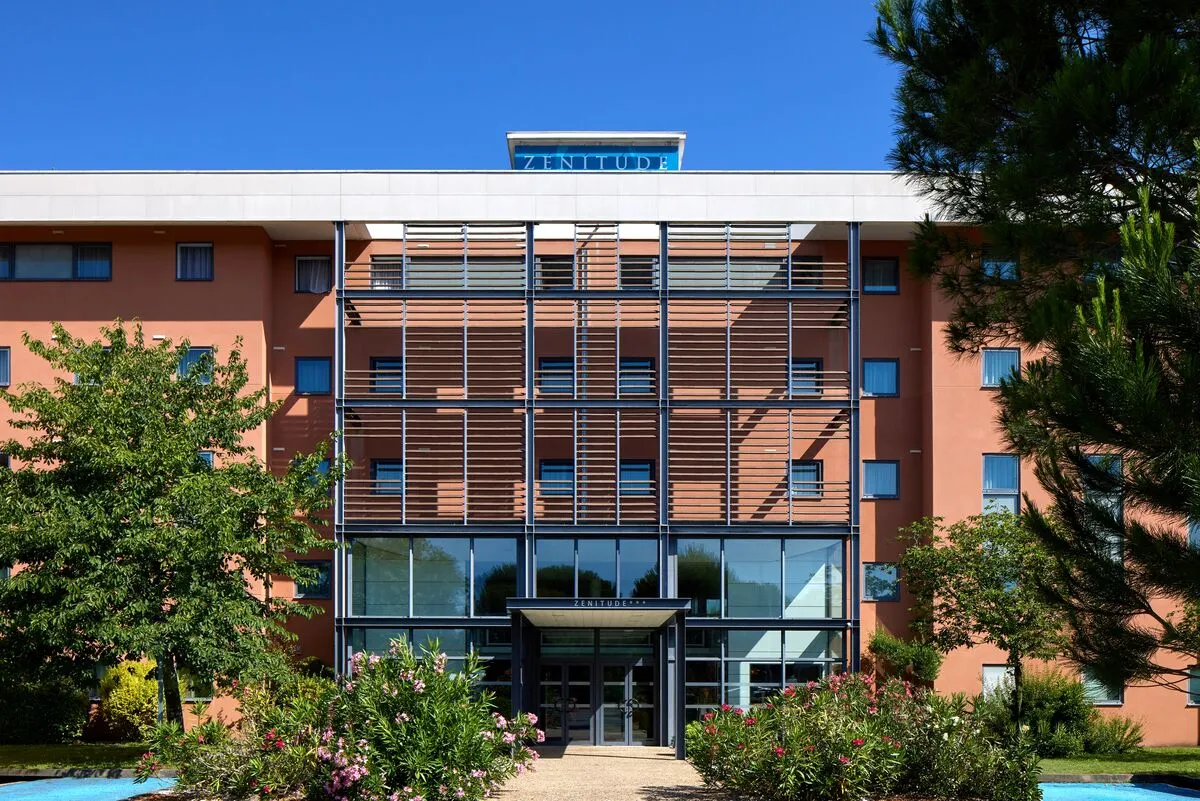 Modern facade of Zenitude Bordeaux Airport hotel featuring terracotta walls, horizontal sun-shading panels, glass entrance, and landscaped surroundings